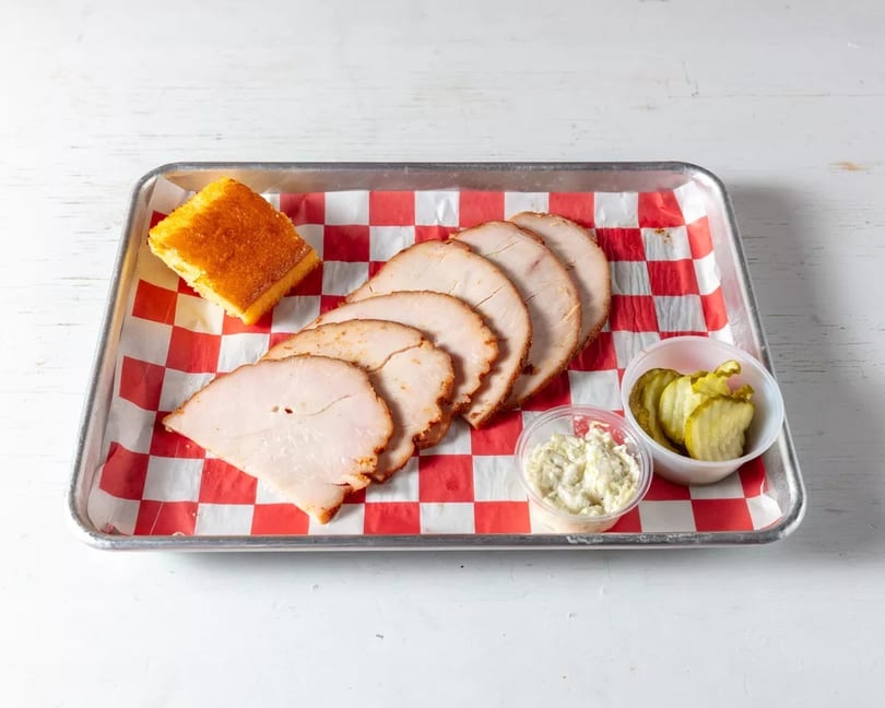 Sliced turkey, cornbread, coleslaw, and pickles on a red and white checkered tray.