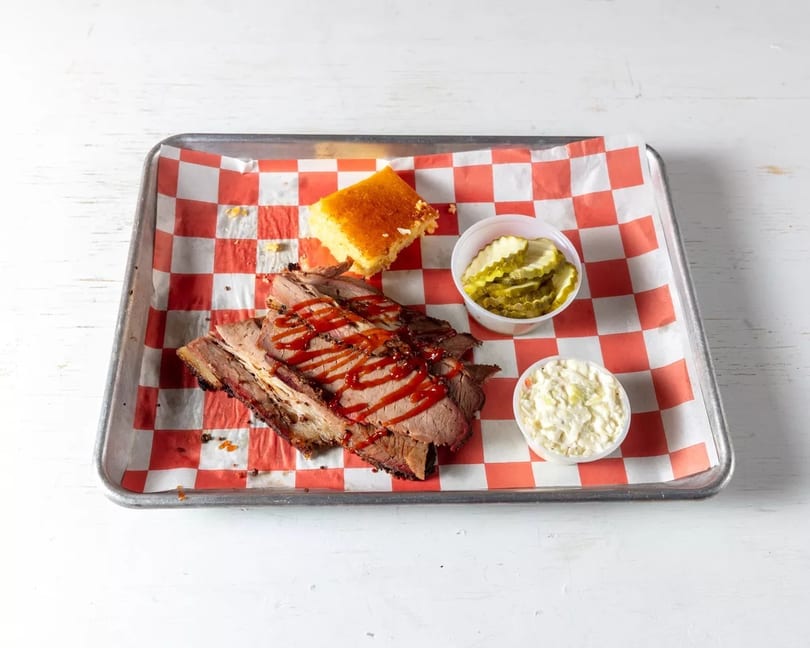 Smoked brisket with BBQ sauce, cornbread, pickles, and coleslaw on a checkered paper-lined tray.