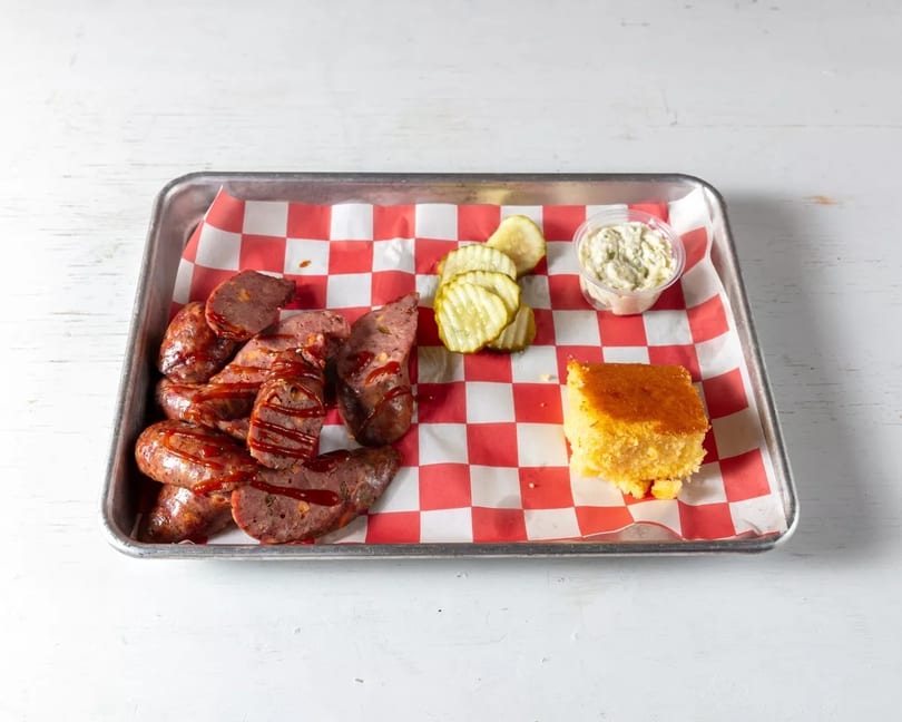 Smoked sausage with BBQ sauce, pickles, cornbread, and a side of tartar sauce on a tray.