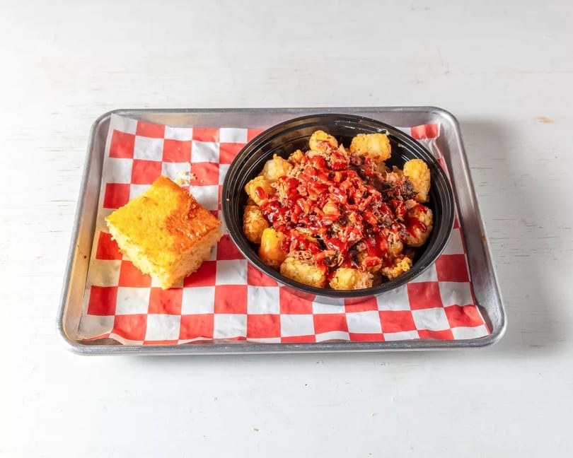 A tray with a bowl of tater tots topped with pulled pork and BBQ sauce, and a piece of cornbread.