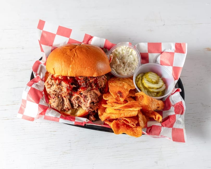 Pulled pork sandwich with barbecue sauce, chips, coleslaw, and pickles in a basket.