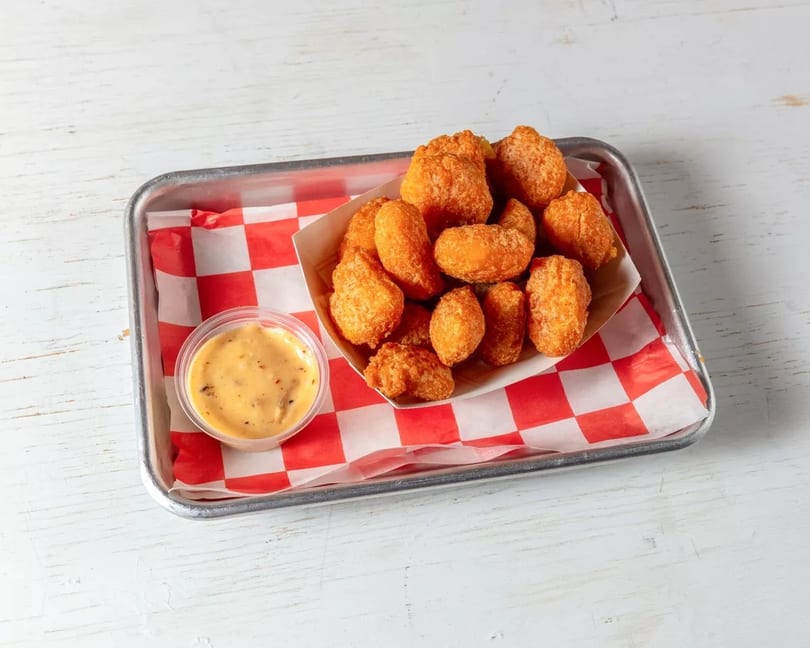 Golden fried tater tots in a paper tray with red and white checkered liner, served with dipping sauce.