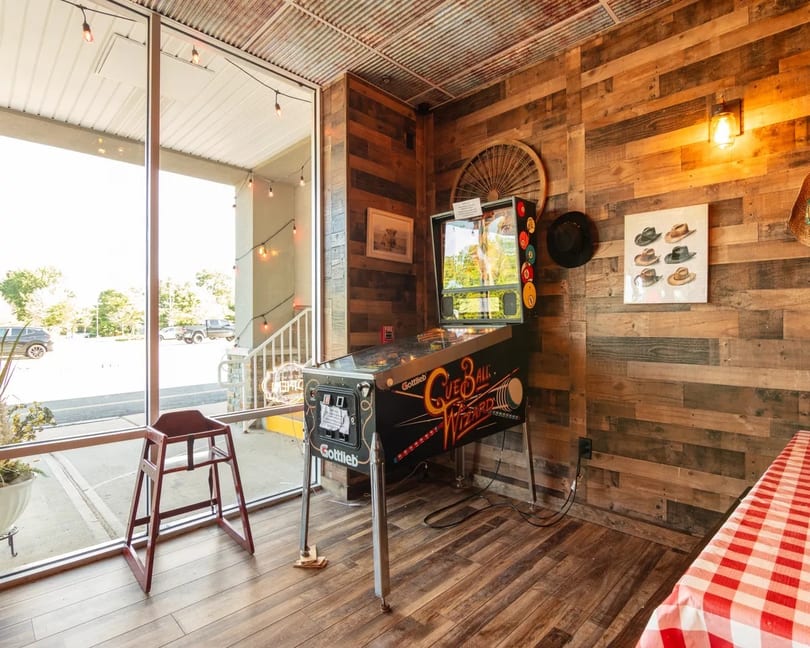 A vintage pinball machine stands next to a large window in a rustic room with wood-paneled walls.