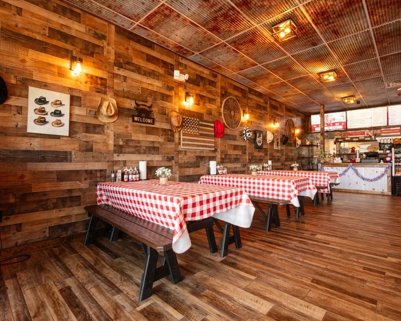 Rustic restaurant interior with wooden walls, red and white checkered tablecloths, and a service counter.