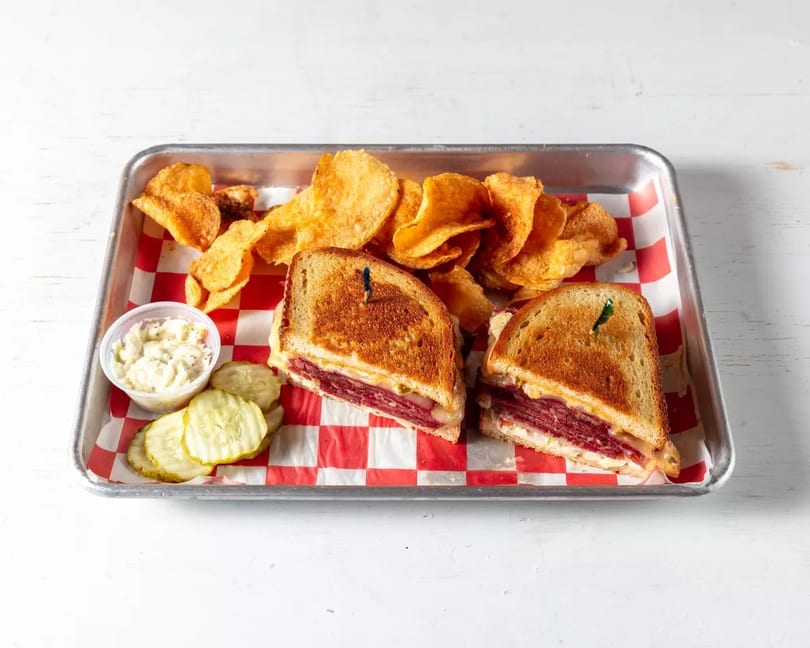 A reuben sandwich cut in half, with potato chips, coleslaw, and pickles on a checkered tray.
