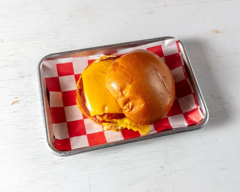 Breakfast sandwich with cheese, egg, and meat on a bun, served on a tray with checkered paper.